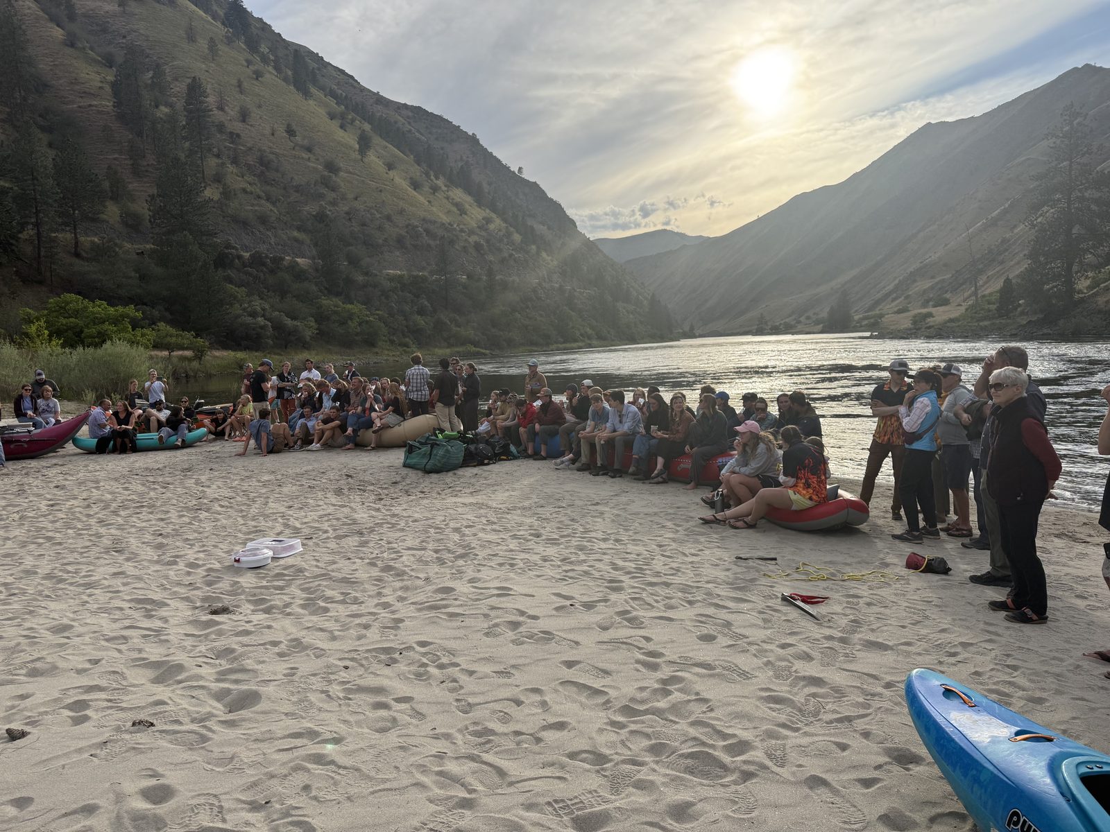 A large gathering on the river beach at golden hour, rafts lined along the shore