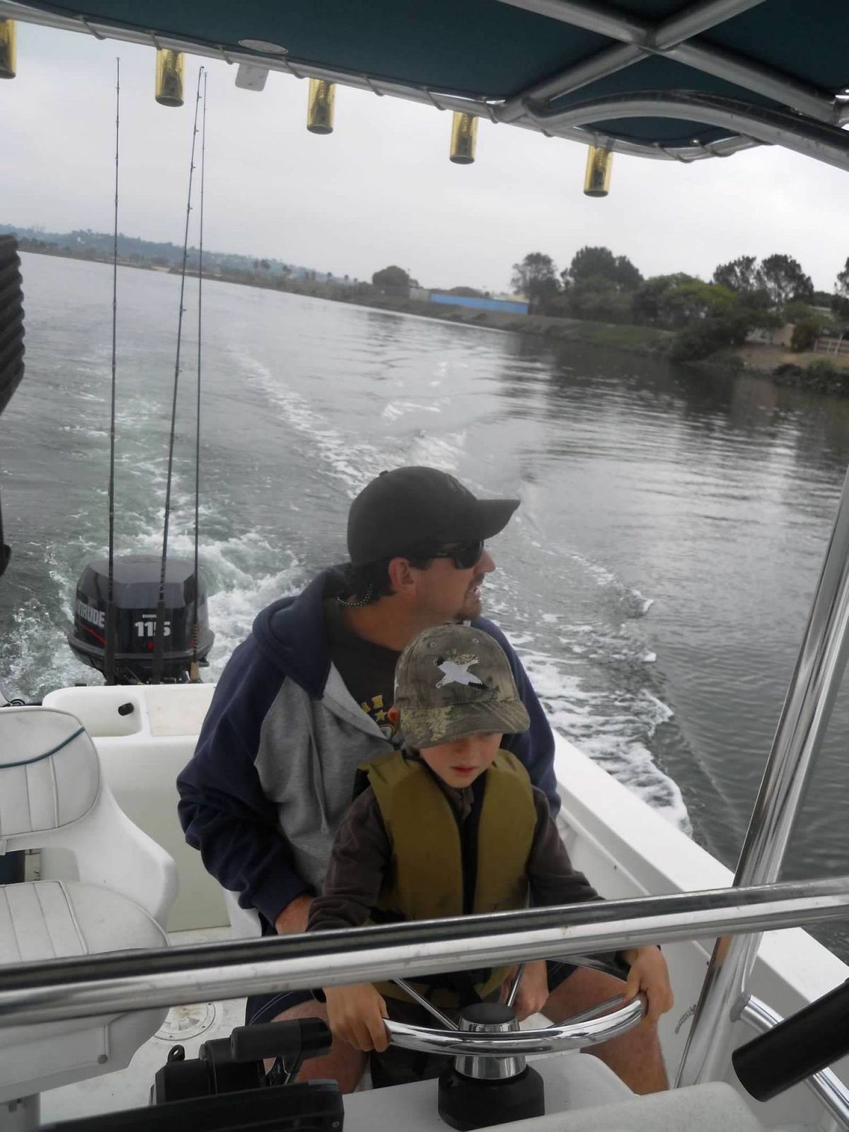 Young Scott driving a boat with his dad behind him, both wearing caps