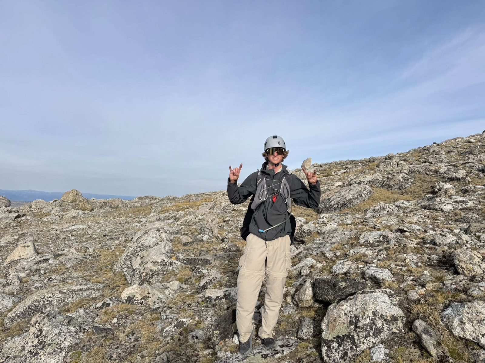 Scott on a rocky ridge, helmet on, both hands throwing horns up, grinning