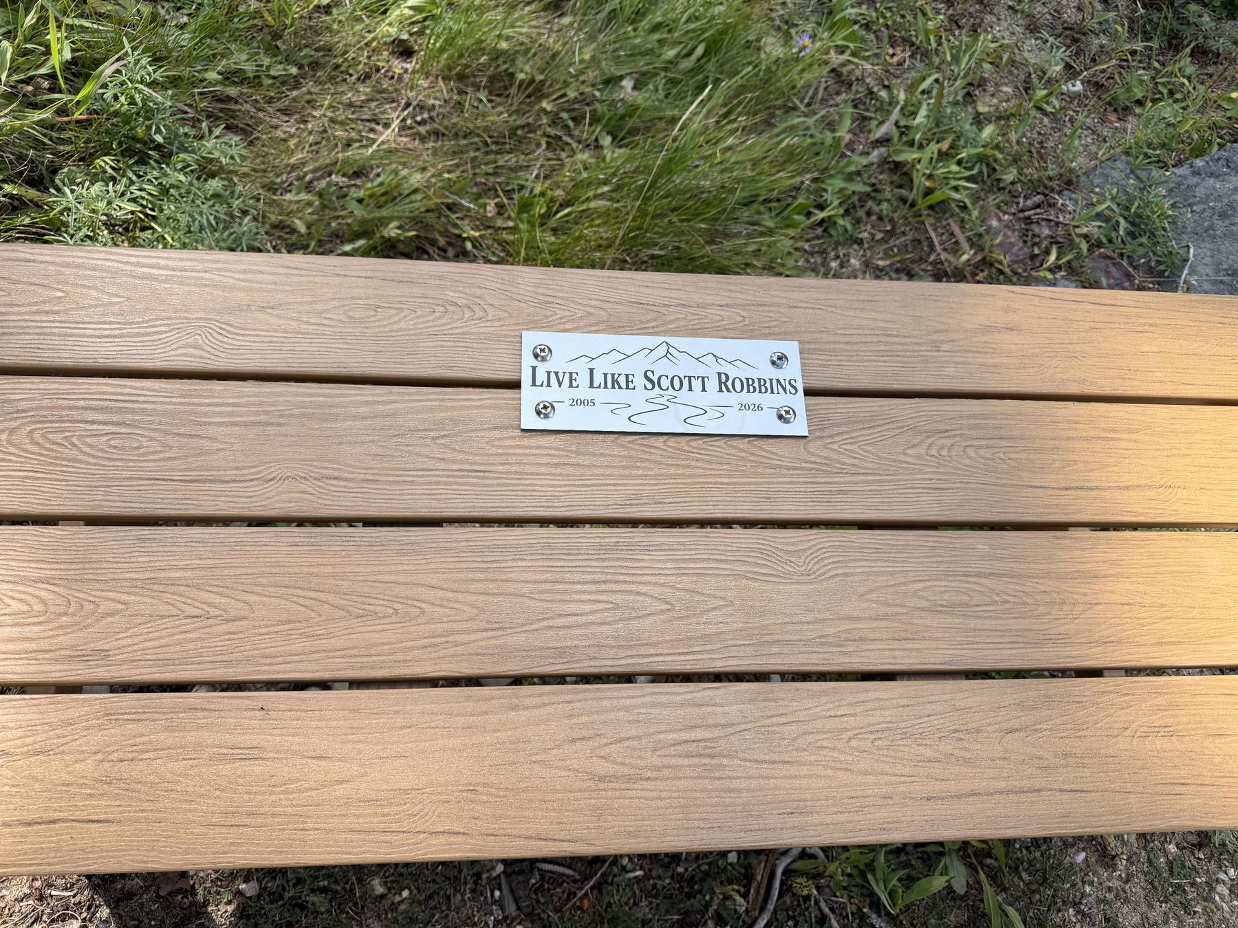 The plaque on the bench &mdash; Live Like Scott Robbins, 2005 to 2026, with mountains engraved above the lettering