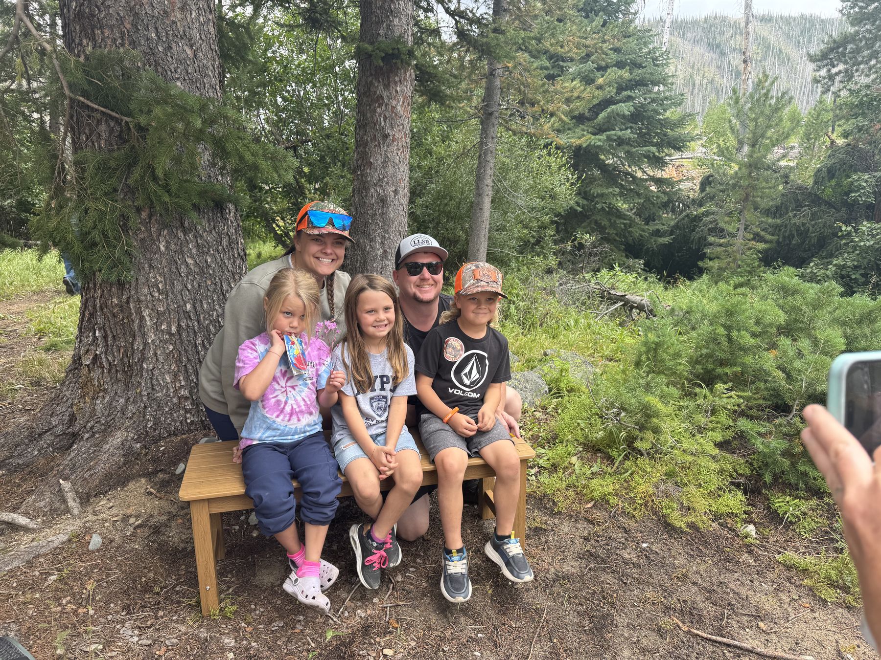 A family sitting together on Scott&rsquo;s bench, three kids between them, under the pines